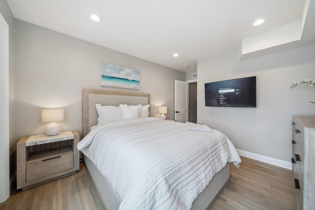 Elegant bedroom with light wood flooring and a light upholstered headboard on a queen bed with white textured bedding. A wall-mounted TV is visible on the right, and a light wood dresser is on the far right.