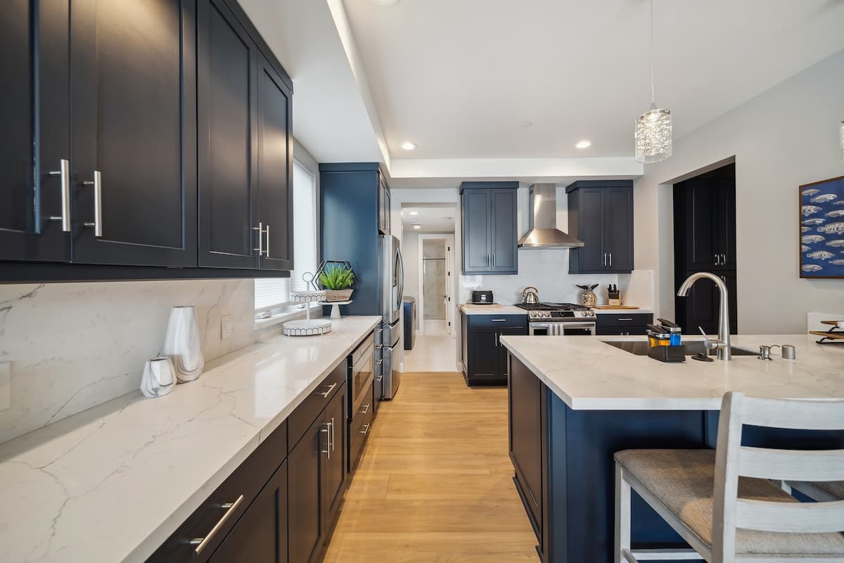 A galley-style kitchen features dark blue shaker cabinets, white marble countertops, and a matching backsplash, extending along both walls. A large island with a sink and bar seating is visible, complemented by stainless steel appliances and light wood flooring.