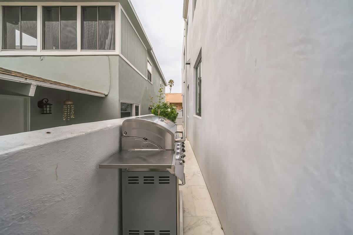 Narrow outdoor walkway between two buildings, featuring a stainless steel BBQ grill, with a glimpse of a neighboring house and lush greenery in the background.