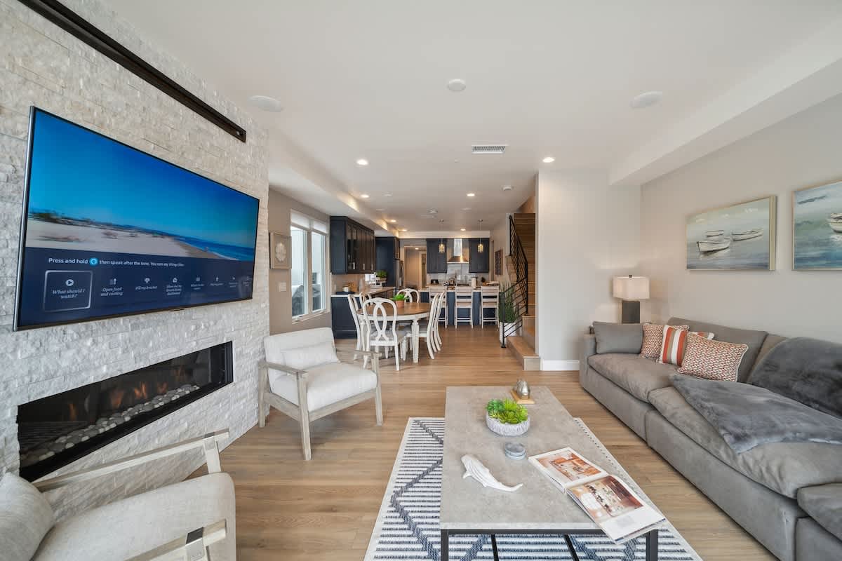 An open-concept living area showcases a grey sectional sofa and armchairs facing a stacked stone fireplace with a mounted TV, flowing into a dining space with a round white table and chairs. The adjacent kitchen with dark blue cabinets is visible, all on light wood flooring.