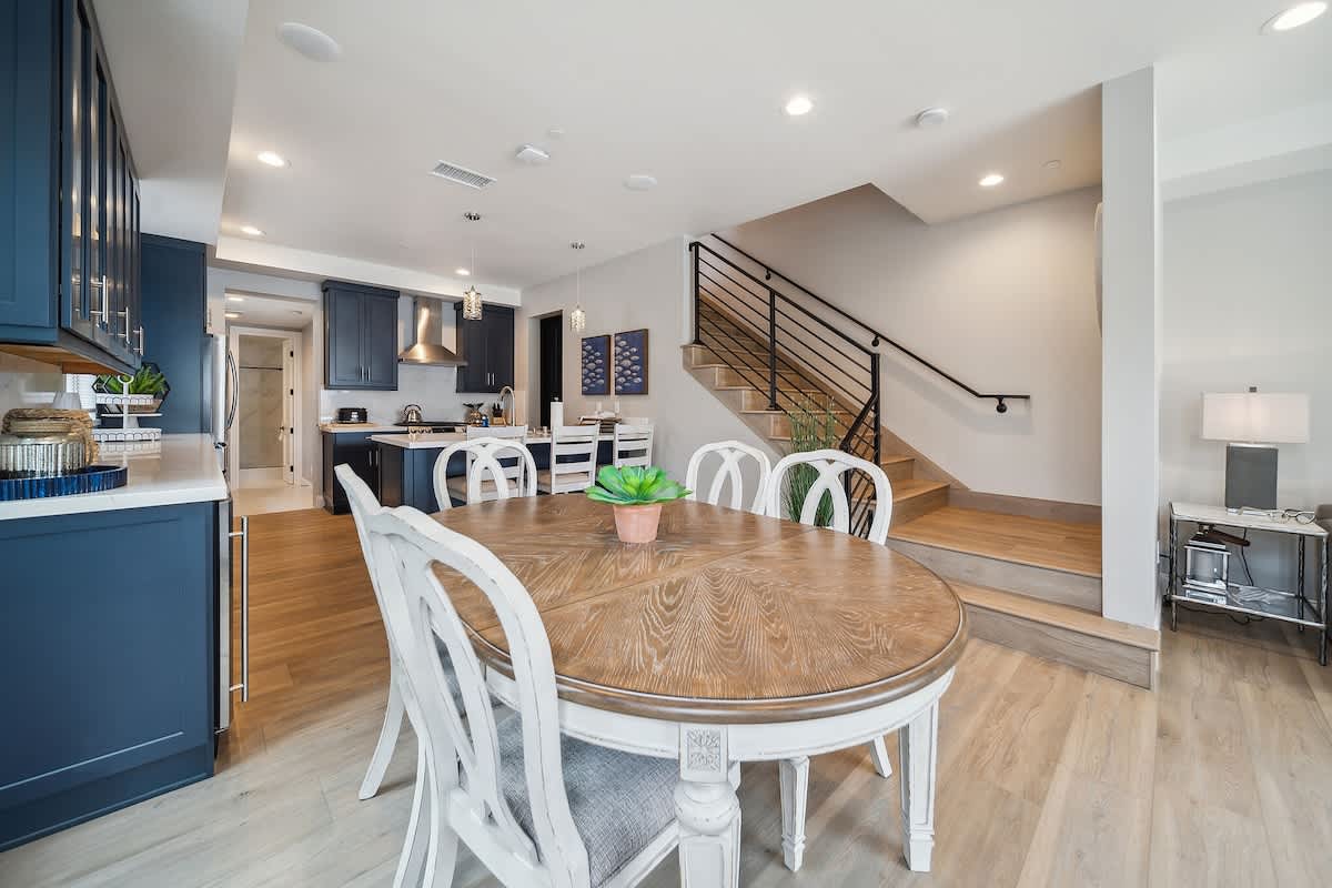 An open-concept dining area features a round wooden table with white chairs, positioned between a modern kitchen with dark blue cabinets and a contemporary staircase with black metal railings. Light wood flooring extends throughout the bright space.