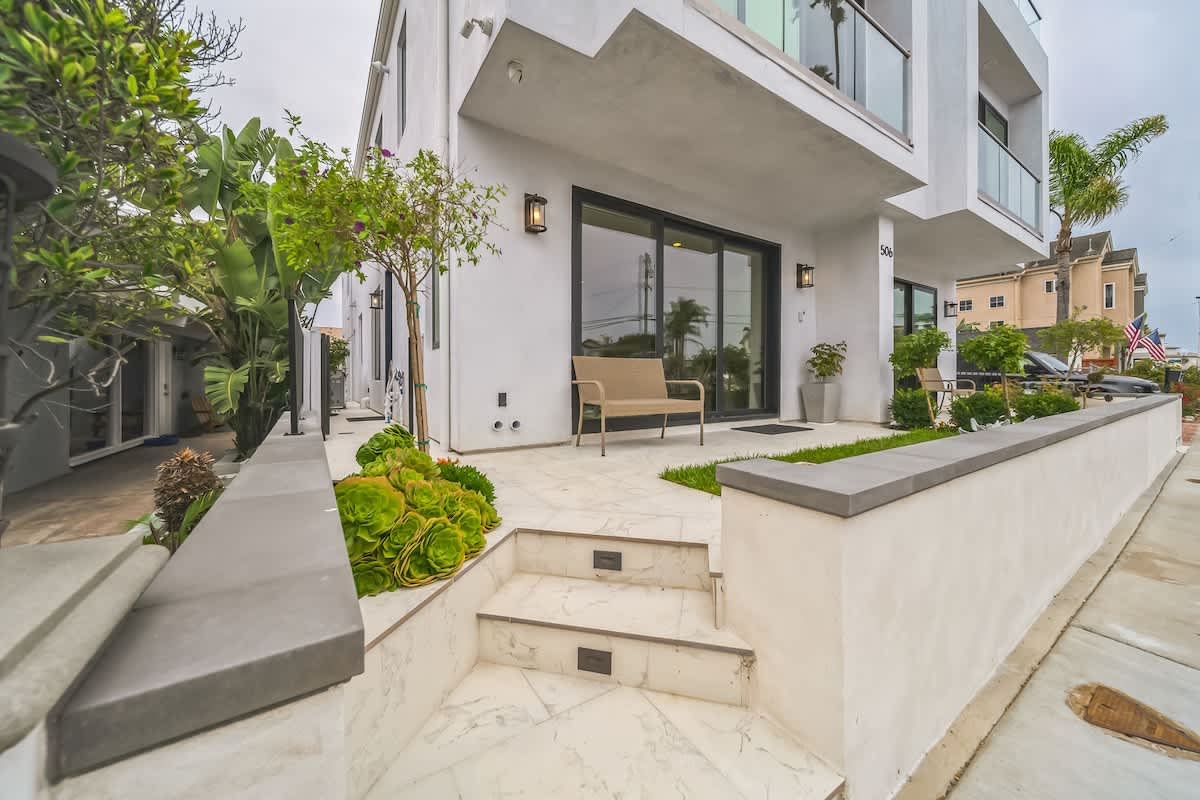 Modern white multi-story building exterior with a clean design, featuring a tiled entryway with steps, lush green landscaping, and a small bench by the entrance.