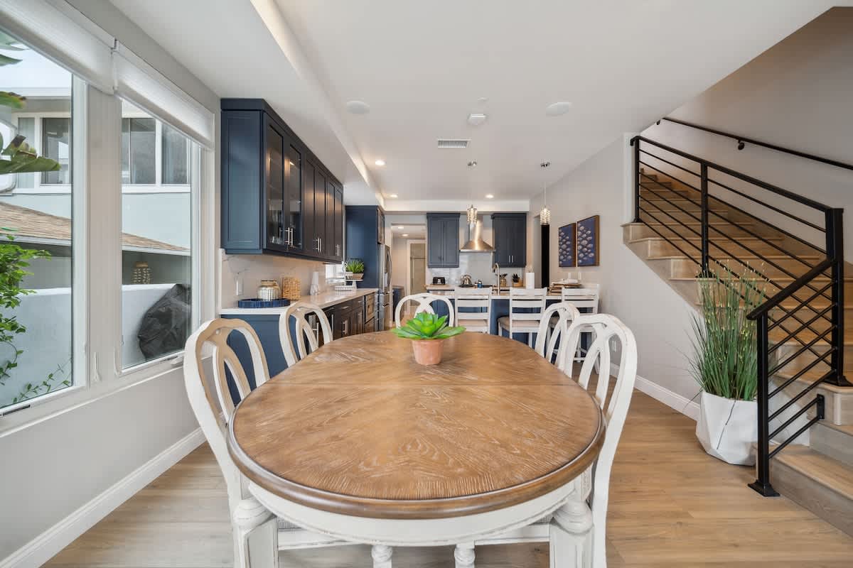 A rustic oval wooden dining table with white chairs is centrally placed in this dining area, adjacent to a kitchen with dark blue cabinets and white countertops. A modern staircase with black railings leads upstairs, and large windows offer views of the neighborhood.