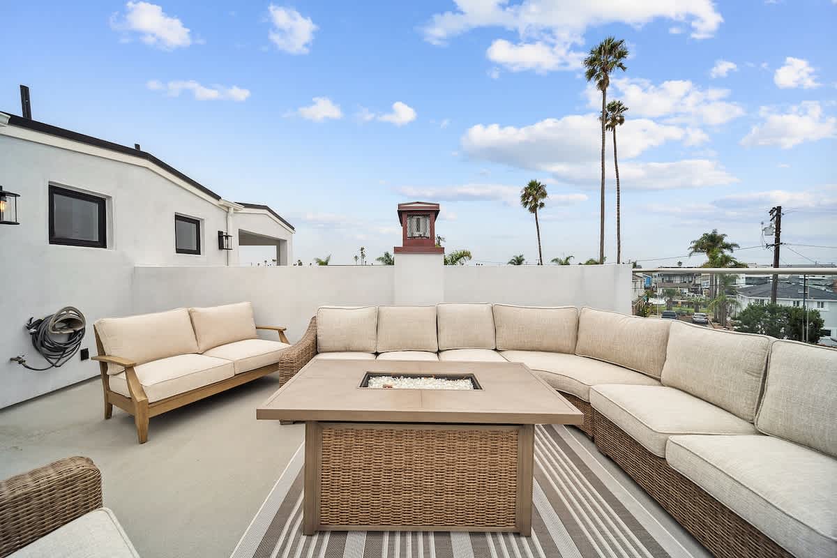 Rooftop deck with a large wicker sectional sofa and a fire pit table, alongside a wooden-framed outdoor sofa, set against a white stucco building with black-framed windows and distant palm trees.