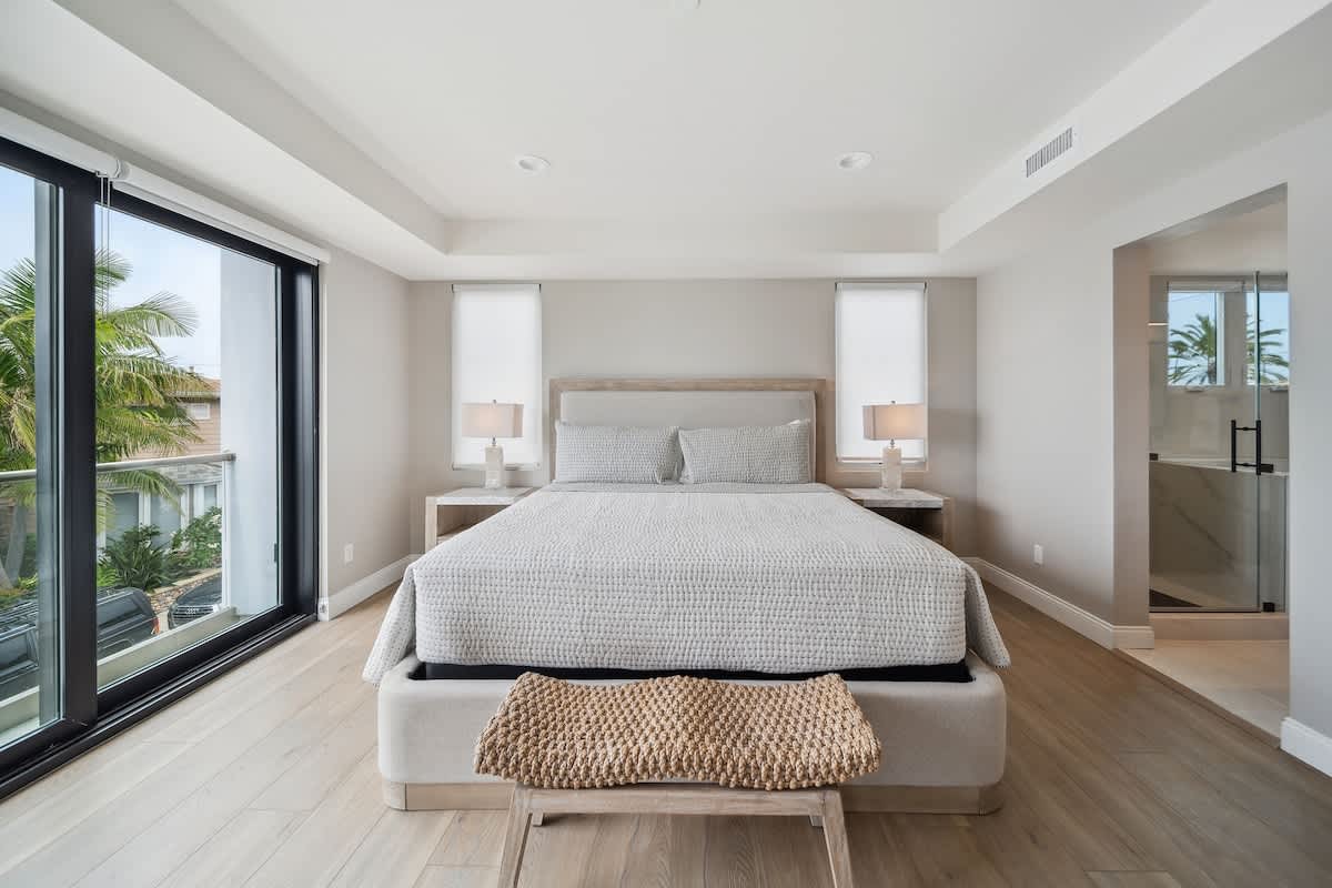 Bright bedroom featuring a large bed with a light gray quilted comforter, flanked by nightstands with lamps, and large sliding glass doors offering views of palm trees and the neighborhood.