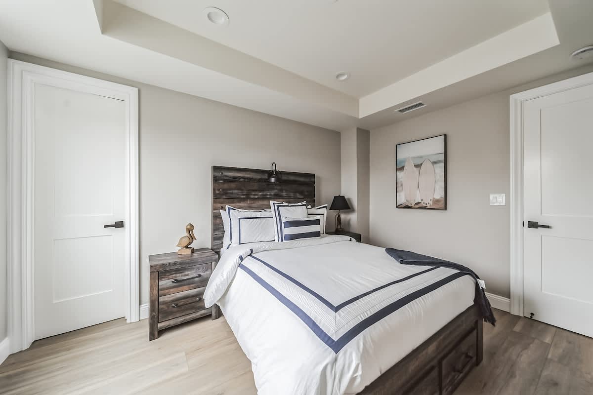 View of a bedroom with light wood flooring and a rustic dark wood headboard, featuring a white bed with blue striped bedding. A nightstand with a lamp and a framed artwork of surfboards are visible, along with two white doors.