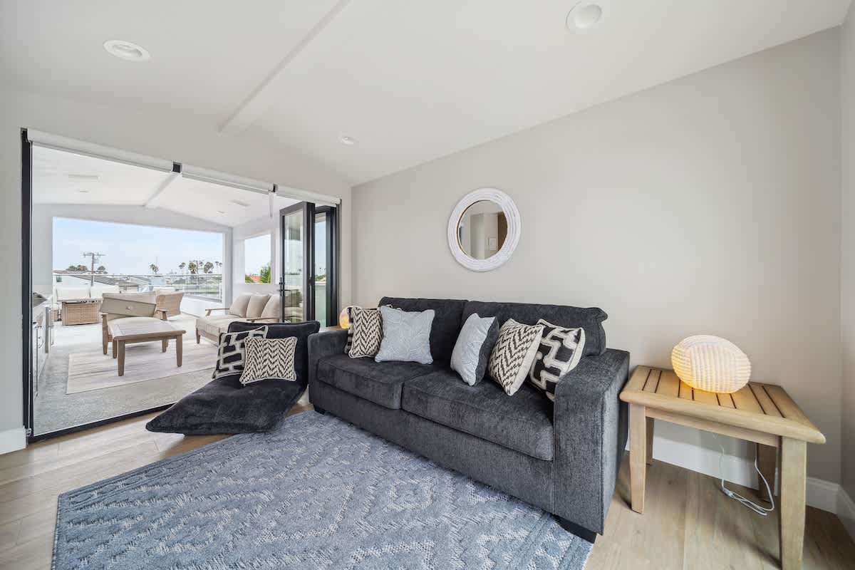 Cozy living room featuring a dark gray sofa adorned with patterned pillows, a light blue geometric rug, and a round white mirror on the wall, with bi-fold doors opening to an outdoor deck.