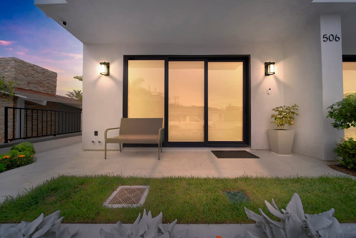 Front exterior of a modern white building at dusk, featuring large sliding glass doors, a small grassy area, and a bench, with warm light reflecting on the glass.