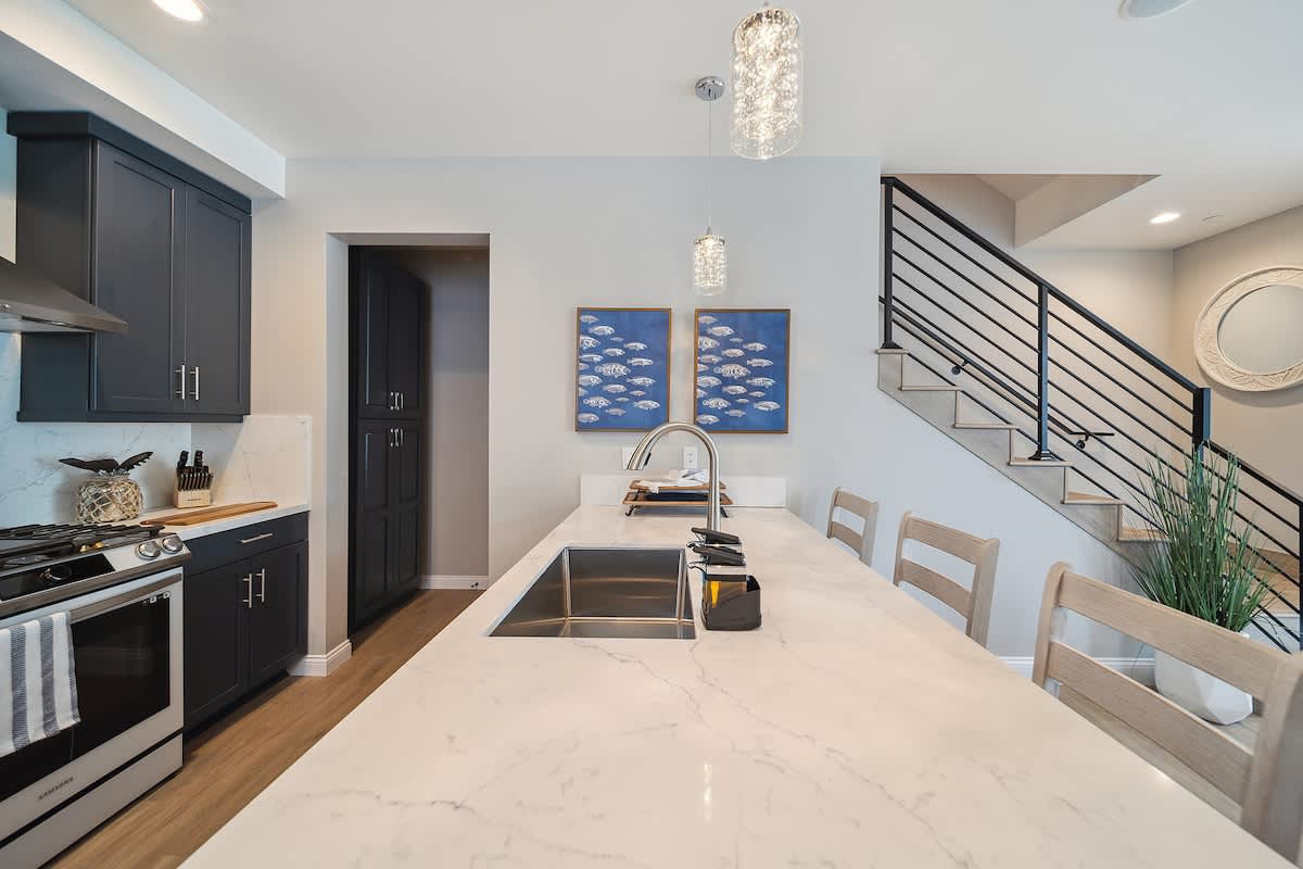 A close-up view of the kitchen island features a stainless steel sink with a modern faucet, set into white quartz countertops. Two crystal pendant lights illuminate the space, with blue fish artwork visible on the wall next to a dark pantry door.