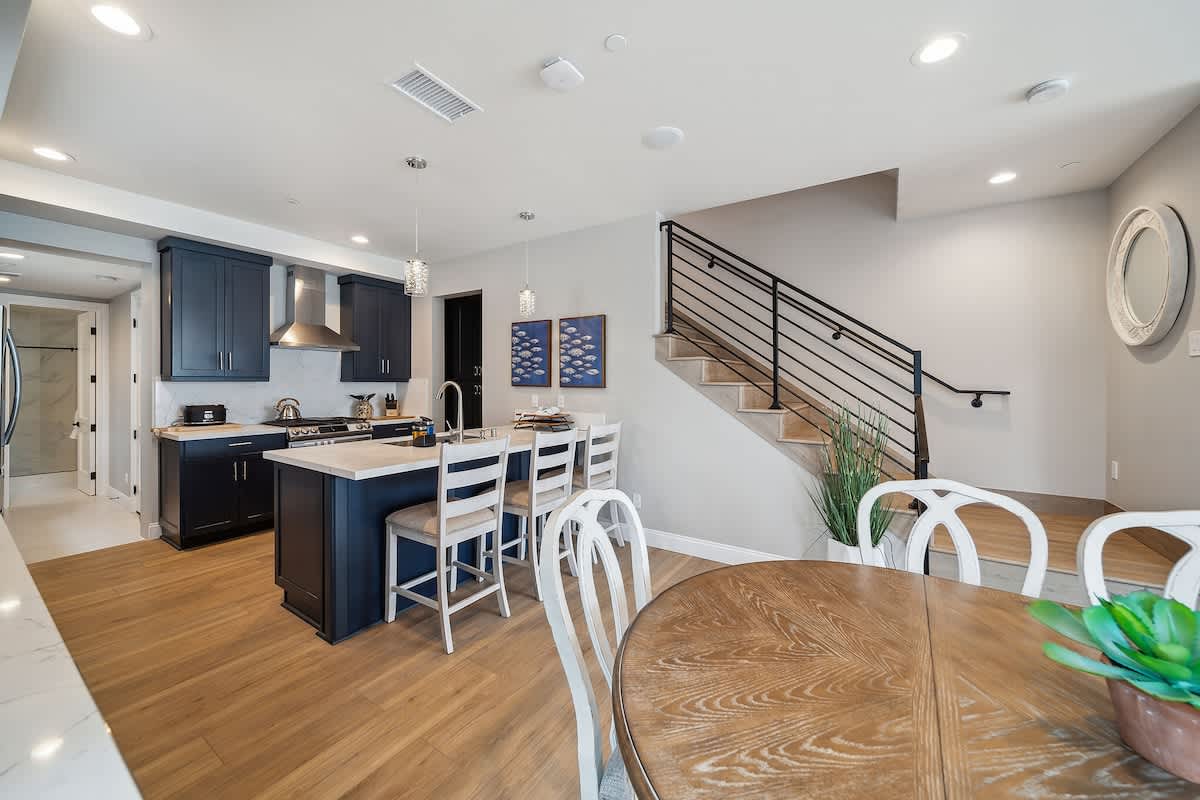 An open-concept view showcases the dining area with a round wooden table and white chairs, the kitchen island with bar stools, and a modern staircase with black metal railings. Light wood flooring unifies the bright, contemporary living space.