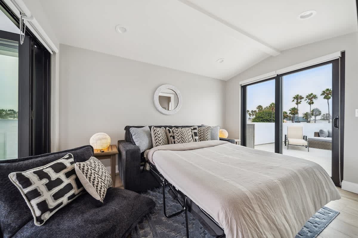 Bright room with a pull-out sofa bed, light wood flooring, and large sliding glass doors leading to an outdoor space with palm tree views.