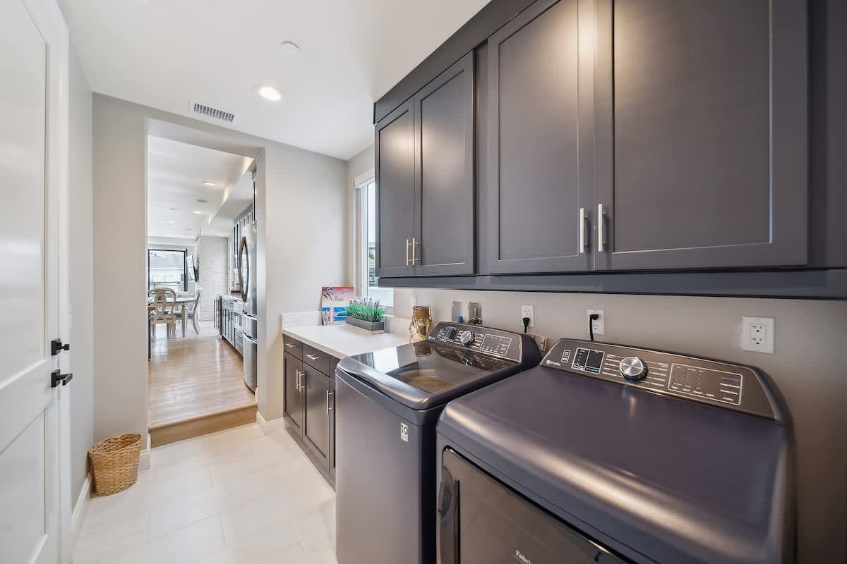 A functional laundry room features dark gray cabinets, white quartz countertops, and a front-loading washer and dryer. The room opens to the main living area with light wood flooring, providing a seamless and integrated space.