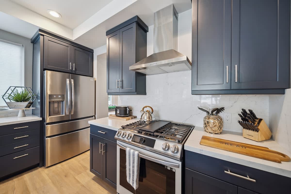 A section of the kitchen highlights a stainless steel French door refrigerator, a gas range with a griddle, and dark blue shaker cabinets with white quartz countertops and a matching backsplash. Kitchen utensils and a toaster are neatly arranged on the counter.