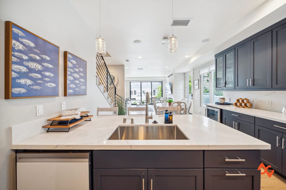 A wide view of the kitchen island with a stainless steel sink, looking towards the dining area and living room with large windows. Dark blue cabinets line the walls, and two crystal pendant lights hang elegantly above the island.