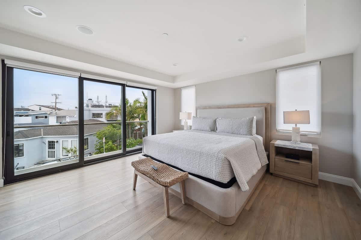 Modern bedroom with a large bed, light wood flooring, and floor-to-ceiling glass doors opening to a balcony with neighborhood views. A woven bench sits at the foot of the bed.