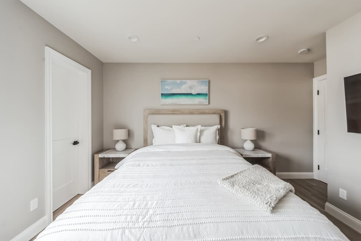 Serene bedroom with light wood flooring and a light upholstered headboard on a queen bed with white textured bedding. Two nightstands with white lamps flank the bed, and a seascape artwork hangs above.
