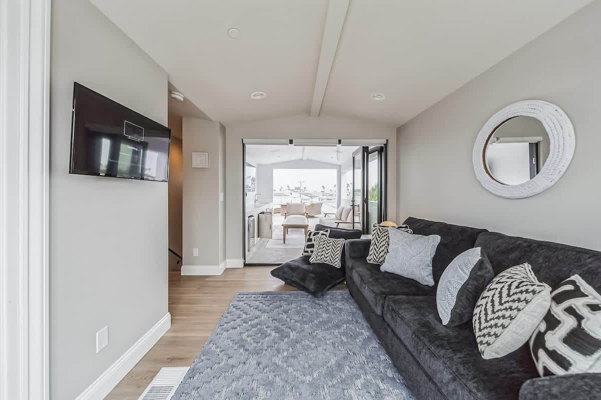 Inviting living room with a dark gray sofa, a light blue geometric rug, and a wall-mounted flat-screen TV, offering a glimpse of an outdoor deck through open bi-fold doors.