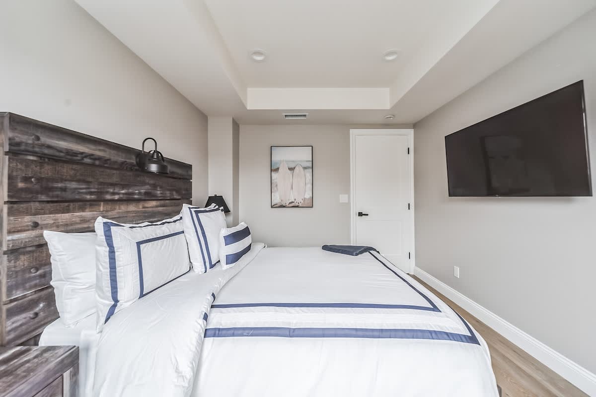 Modern bedroom with light wood flooring and a rustic dark wood headboard, featuring a white bed with blue striped bedding. A large wall-mounted TV is positioned opposite the bed, and a framed surfboard artwork hangs on the wall.