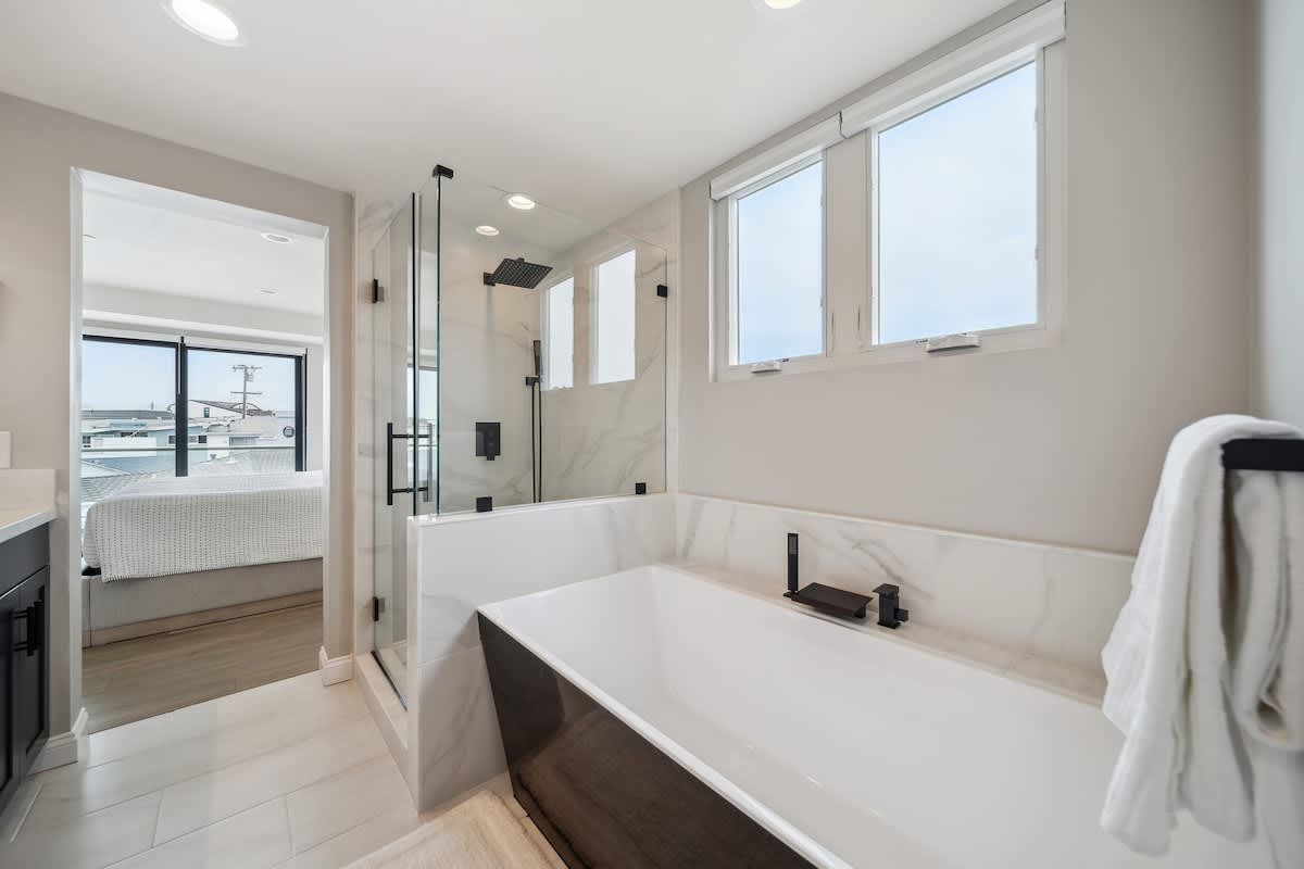 Bright bathroom with a freestanding soaking tub, a glass-enclosed shower with a rain showerhead, and a view into the adjacent bedroom through an open doorway.