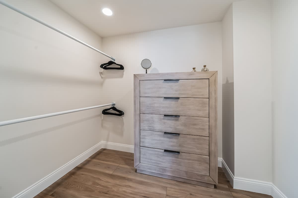 Walk-in closet with light wood flooring, white walls, and two hanging rods, featuring a light wood dresser with dark handles.