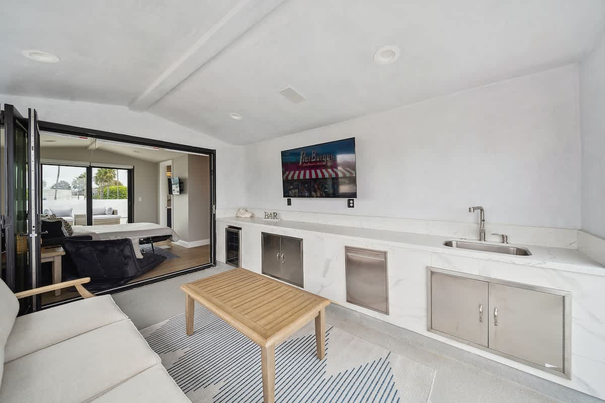 Modern indoor lounge area with a white marble-topped bar featuring a sink, stainless steel appliances, and a wall-mounted TV, opening via bi-fold doors to an outdoor patio with seating.