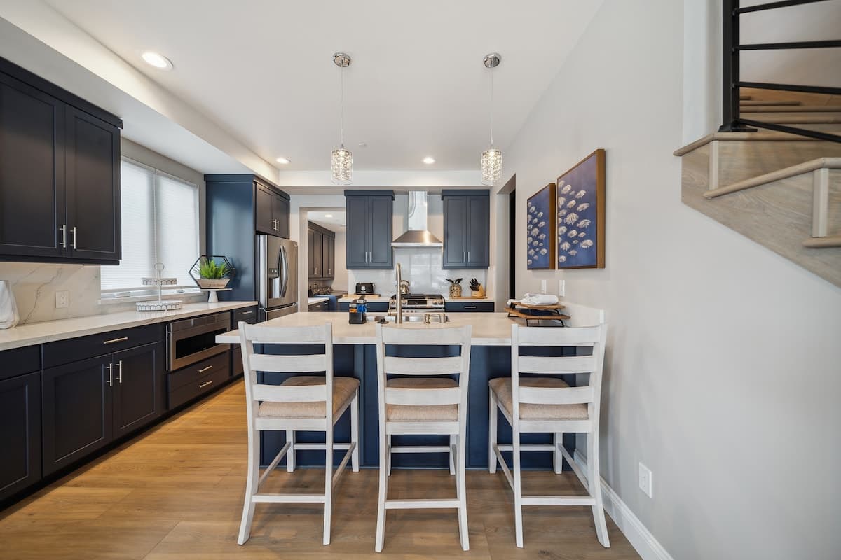 A modern kitchen showcases dark blue shaker-style cabinets, white quartz countertops, and a large island with a stainless steel sink and three white bar stools. Two decorative pendant lights hang above the island, complementing the blue fish-themed artwork.