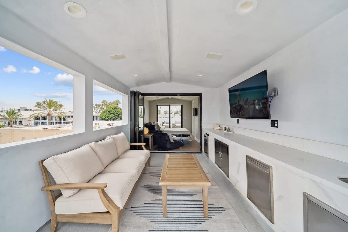 Covered outdoor balcony with a comfortable sofa, coffee table, built-in bar area with a mounted TV, and views of palm trees and the neighborhood.