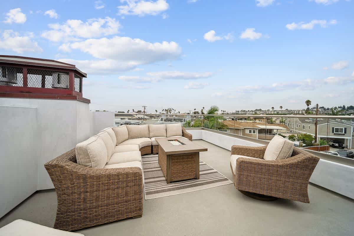 Wide shot of a spacious rooftop deck furnished with a large wicker sectional sofa and a fire pit table, providing sweeping views of the coastal neighborhood and distant ocean through a glass railing.