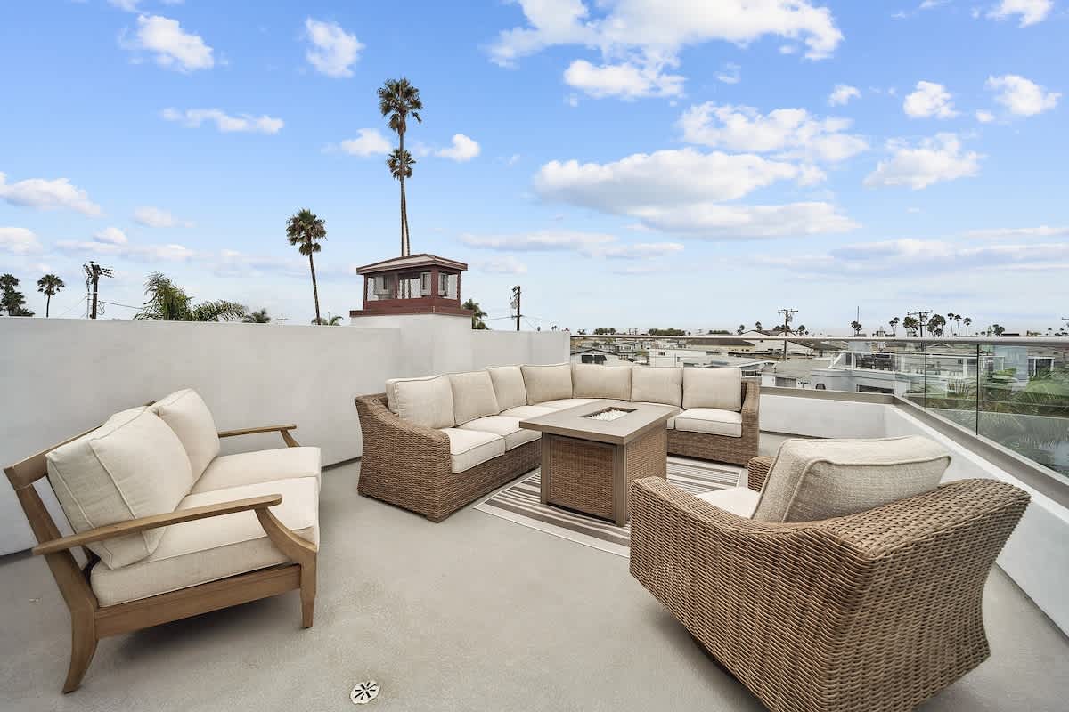 Expansive rooftop deck featuring a large wicker sectional sofa and armchairs with cream cushions, centered around a fire pit table, offering panoramic views of the surrounding neighborhood and palm trees under a blue sky.