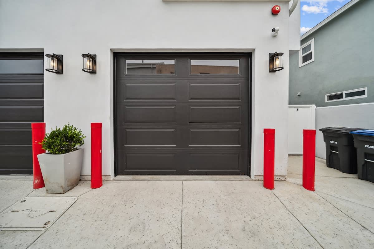 Dark gray paneled garage door with windows, flanked by two outdoor sconces and red bollards, set against a white building facade.