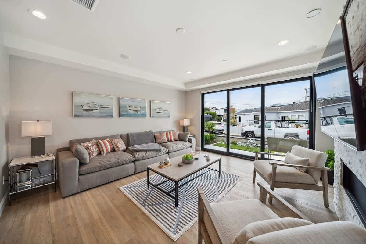 A spacious living room features a long grey sectional sofa adorned with throw pillows, facing a modern coffee table on a geometric striped rug. Large sliding glass doors provide ample natural light and views of the street, while light wood flooring extends throughout the space.