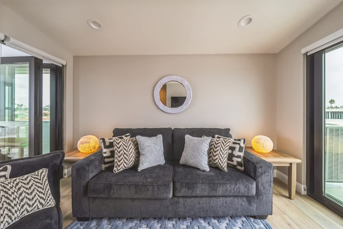 Bright living room featuring a dark gray sofa with patterned pillows, flanked by wooden side tables with spherical lamps, and large glass doors leading to an outdoor space.