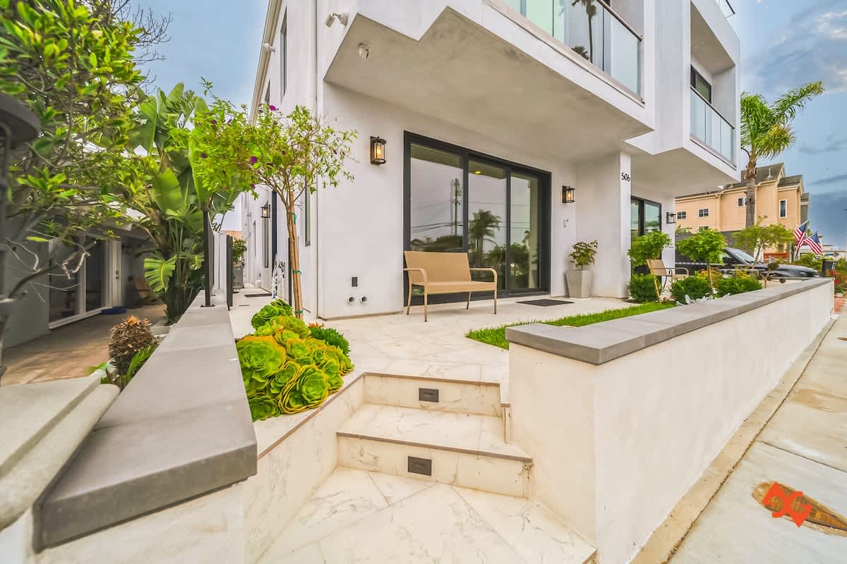Modern white multi-story building exterior with a clean design, featuring a tiled entryway with steps, lush green landscaping, and a small bench by the entrance.