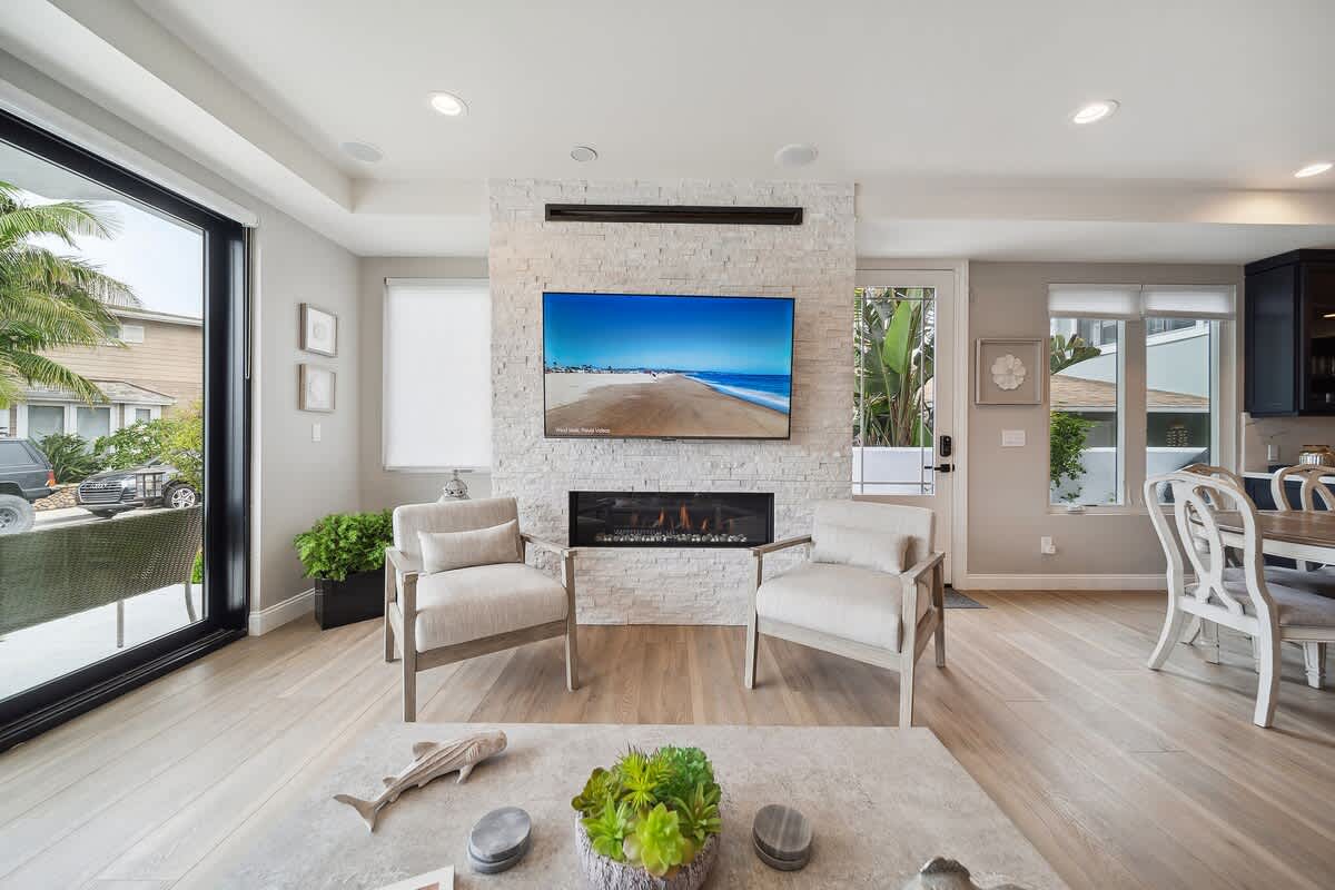 A cozy living room features a modern linear fireplace set into a white stacked stone accent wall, with a large flat-screen TV mounted above. Two light-colored armchairs face the fireplace, while large sliding glass doors open to an outdoor patio, all on light wood flooring.
