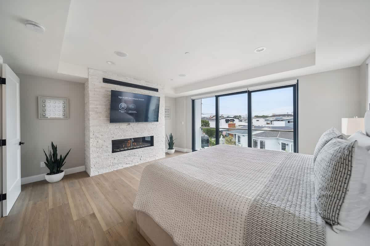 Contemporary bedroom featuring a white stacked stone fireplace with a mounted flat-screen TV, light wood floors, and large sliding glass doors providing outdoor views.