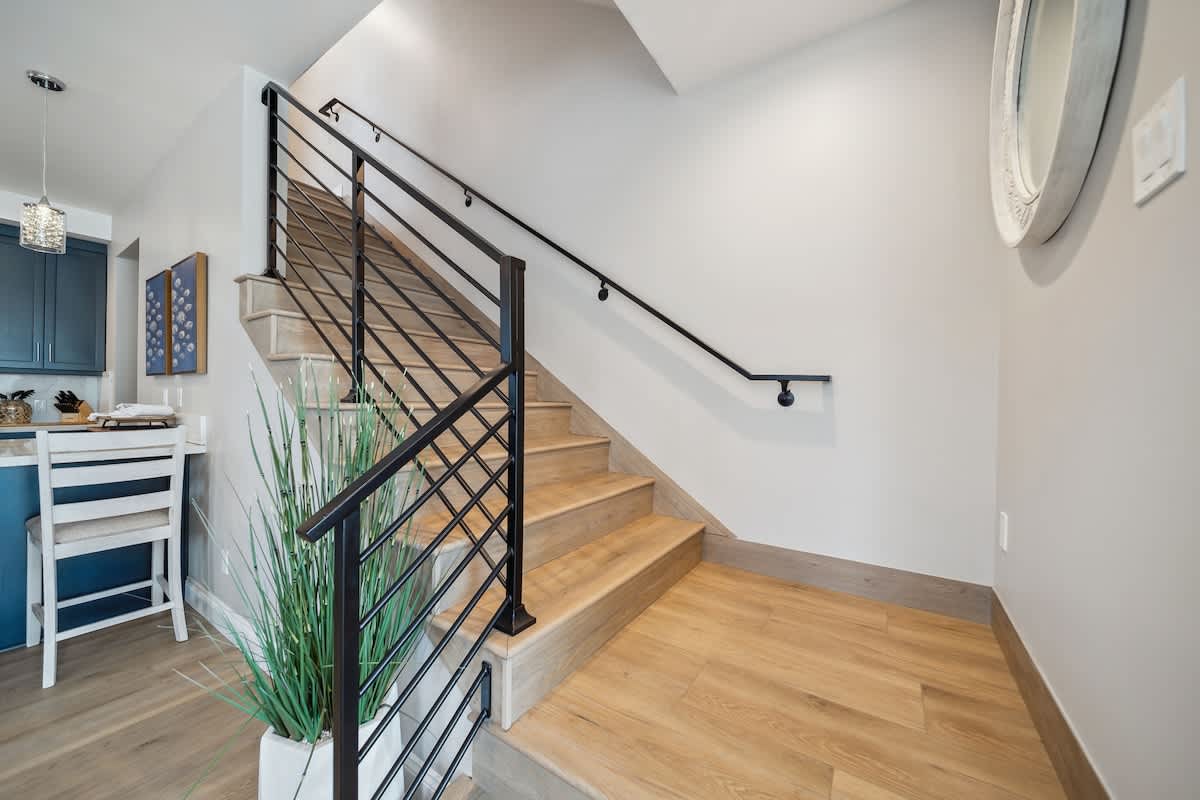 A modern staircase with light wood treads and sleek black metal railings ascends against a light gray wall, featuring a decorative round mirror and a tall potted plant at its base. Light wood flooring continues from the adjacent living areas.