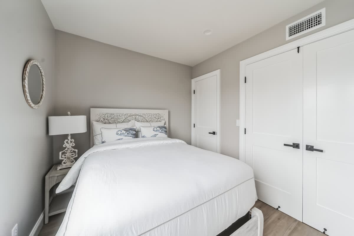 View of a cozy bedroom with a white bed, decorative headboard, and light wood flooring. A white nightstand with a lamp and a round mirror adorn the wall next to the bed, with white closet doors visible.