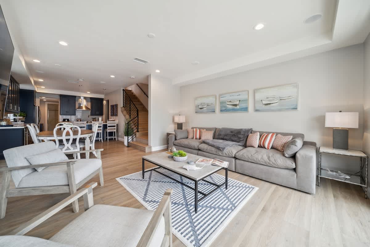 An open-concept living space combines a comfortable grey sectional sofa and armchairs with a dining area and a glimpse of the kitchen. Light wood flooring unifies the space, leading towards a modern staircase with black railings, all under recessed lighting.