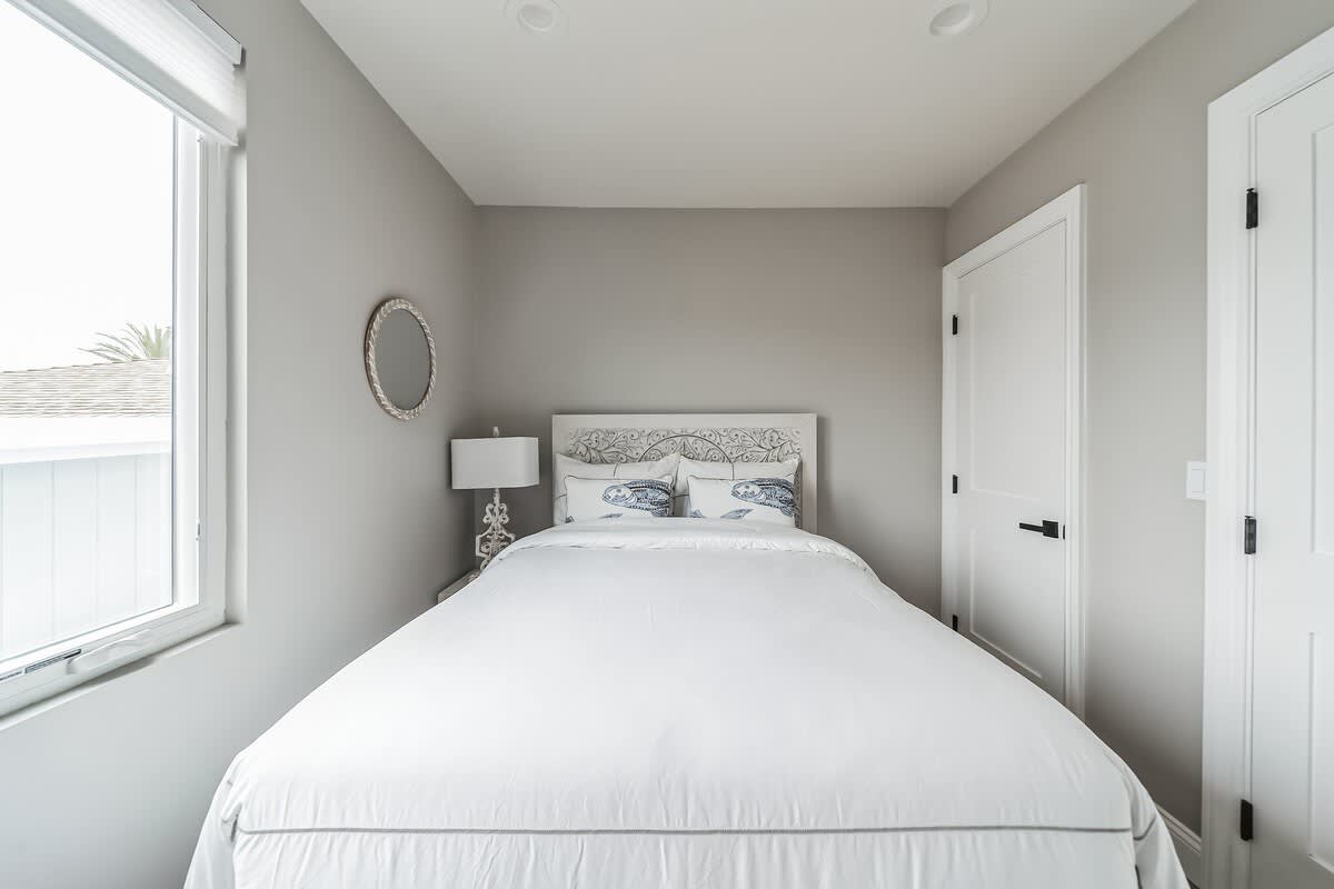 Bright bedroom with light gray walls and wood flooring, featuring a white bed with a decorative headboard. A large window with white blinds overlooks a neighborhood, and white closet doors are visible on the right.