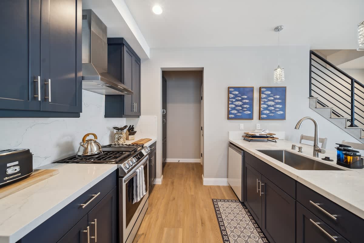 This galley-style kitchen features dark blue cabinets, white quartz countertops, and a stainless steel gas range with a matching range hood. Light wood flooring runs through the space, leading to a dark pantry door and an open living area.