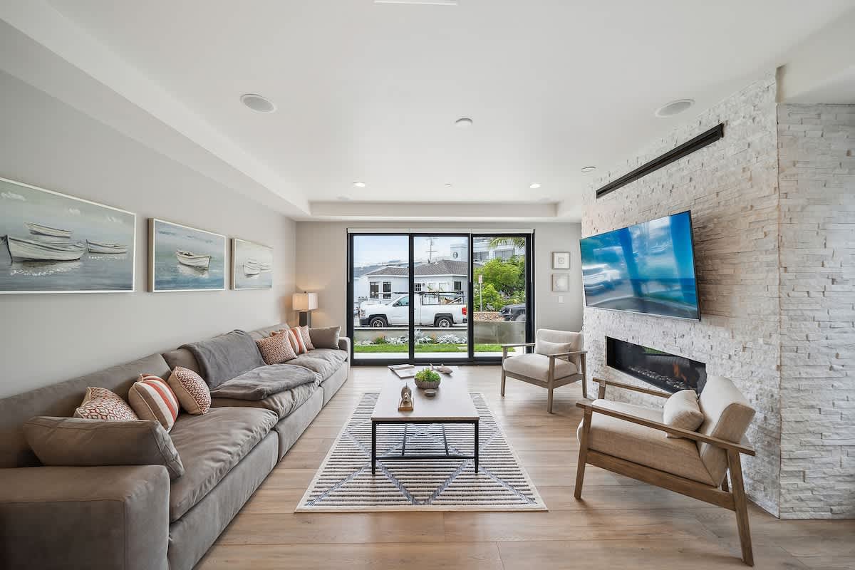 A bright and airy living room features a long grey sectional sofa and two light-colored armchairs arranged around a modern coffee table on a striped rug. A white stacked stone fireplace with a mounted TV anchors one wall, while large sliding glass doors offer views to the outside.