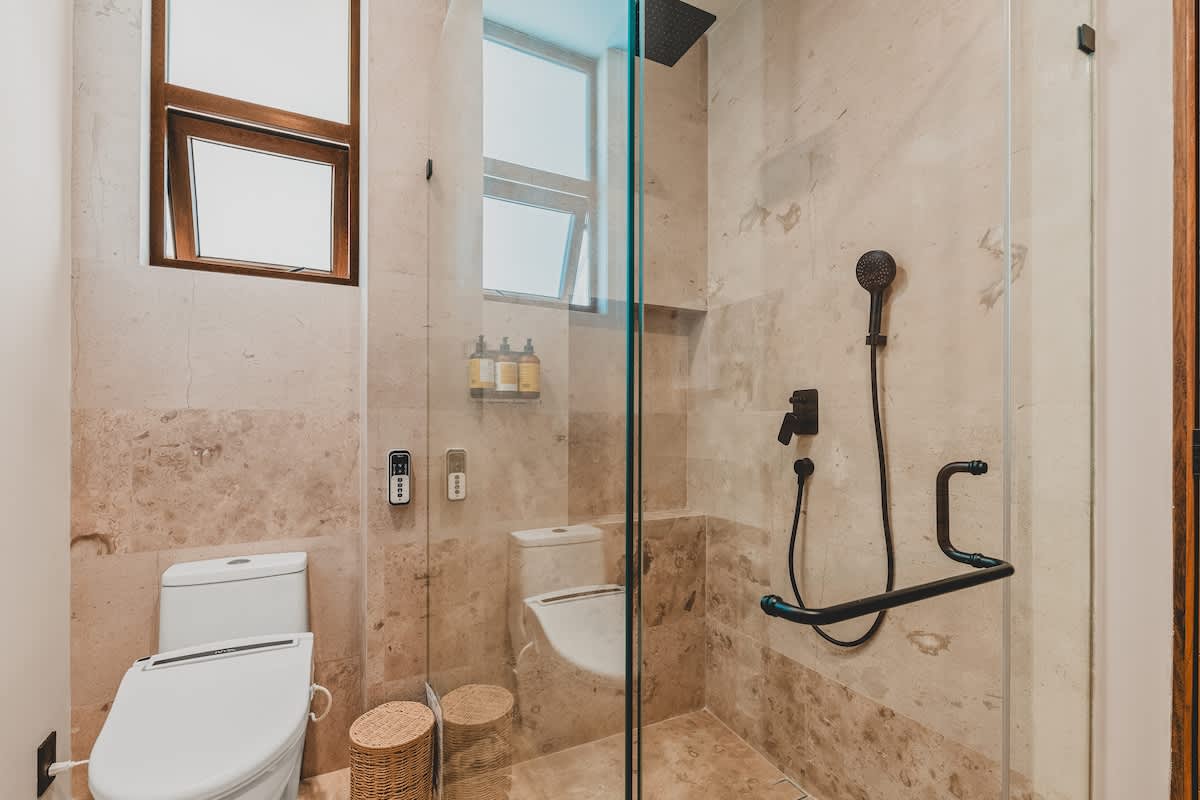 A contemporary shower area with light-colored stone tiles, a glass enclosure, and a black rainfall showerhead with a handheld sprayer. A modern toilet is visible next to the shower, with a small window above.