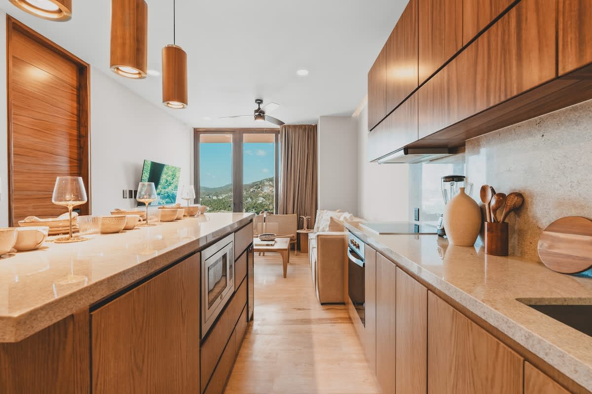 Open-concept kitchen and living area featuring light wood cabinetry, a long dining island, and a comfortable living space with a sofa and chairs, all overlooking a scenic outdoor view through large glass doors.