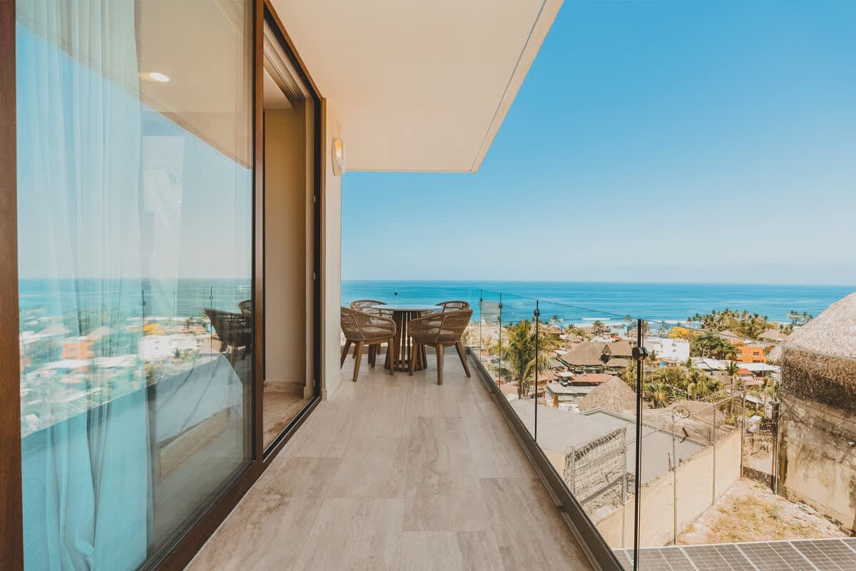 Expansive balcony with a round outdoor dining table and four woven chairs, offering panoramic views of the deep blue ocean and a vibrant coastal town below.