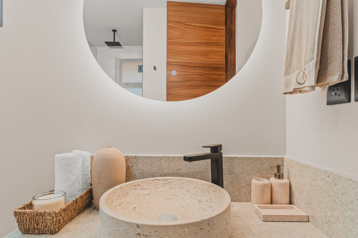 A close-up of the bathroom vanity showcasing a textured stone vessel sink, a sleek black faucet, and a large circular backlit mirror. Decorative items and neatly folded towels are arranged on the light stone countertop.