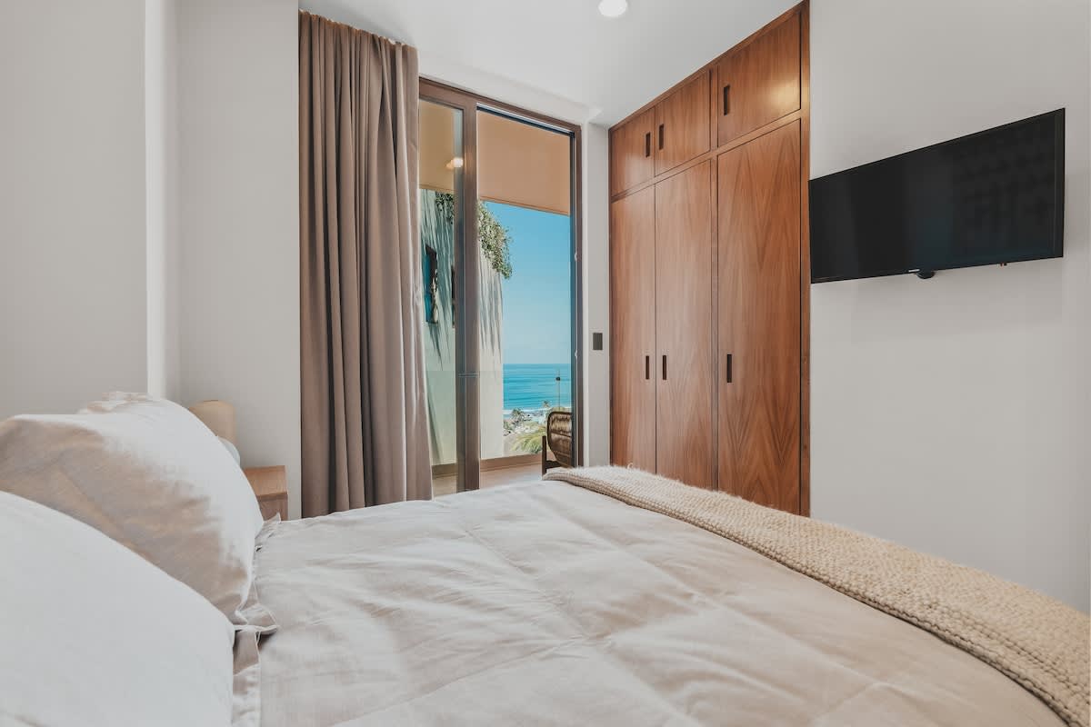 Bright bedroom featuring a large bed with light bedding, a dark wooden built-in wardrobe, and a wall-mounted TV, with sliding glass doors opening to a balcony offering a stunning ocean view.