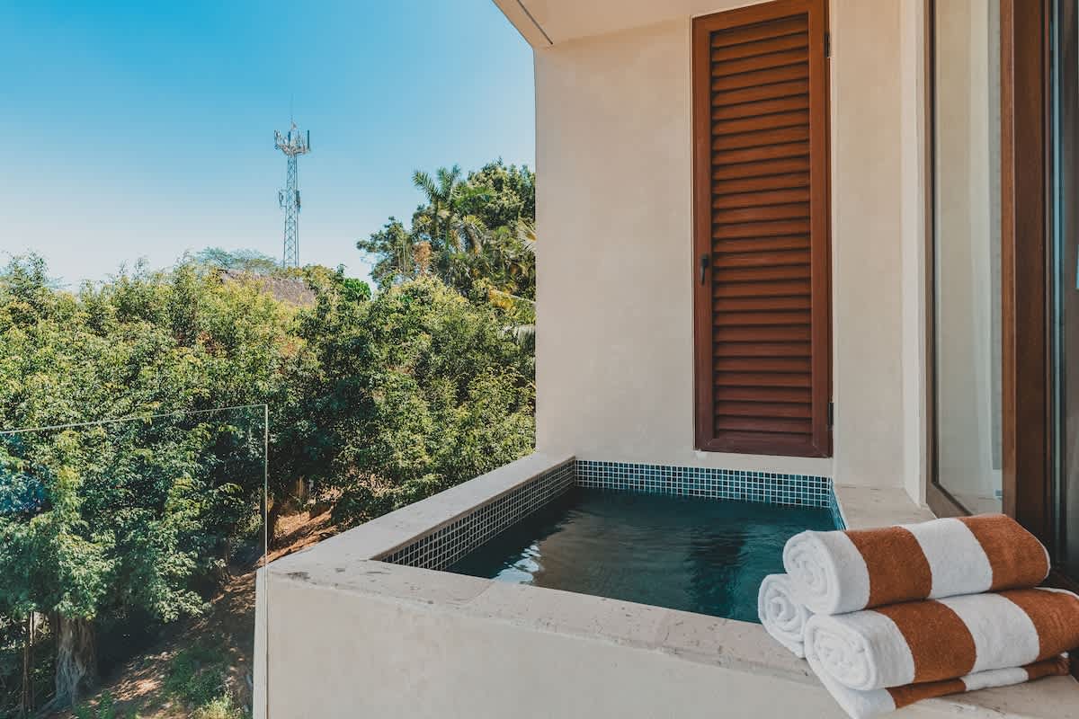 Close-up view of the compact plunge pool on a balcony, featuring blue mosaic tiles and neatly folded white and brown striped towels, with a distant cell tower against a clear sky.