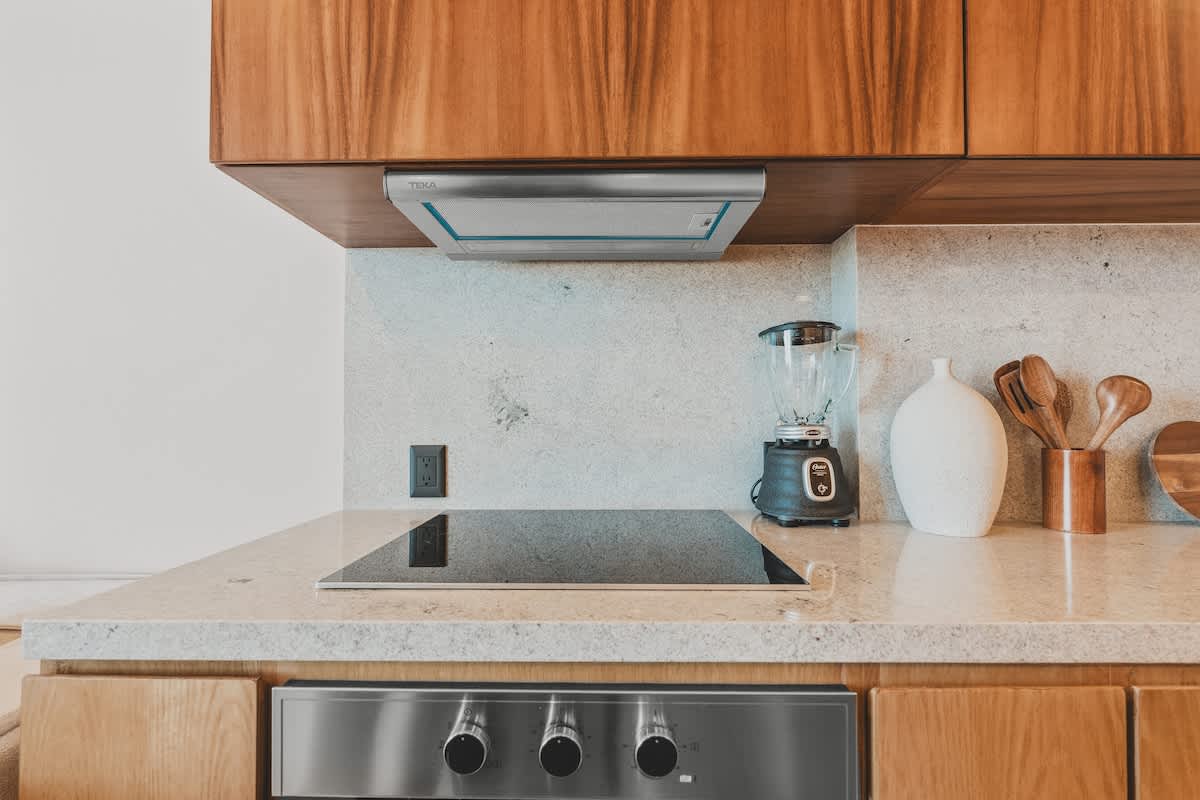 Detailed view of the kitchen countertop with a sleek induction cooktop, a built-in range hood, and a blender, set against a light speckled backsplash and warm wood cabinetry.