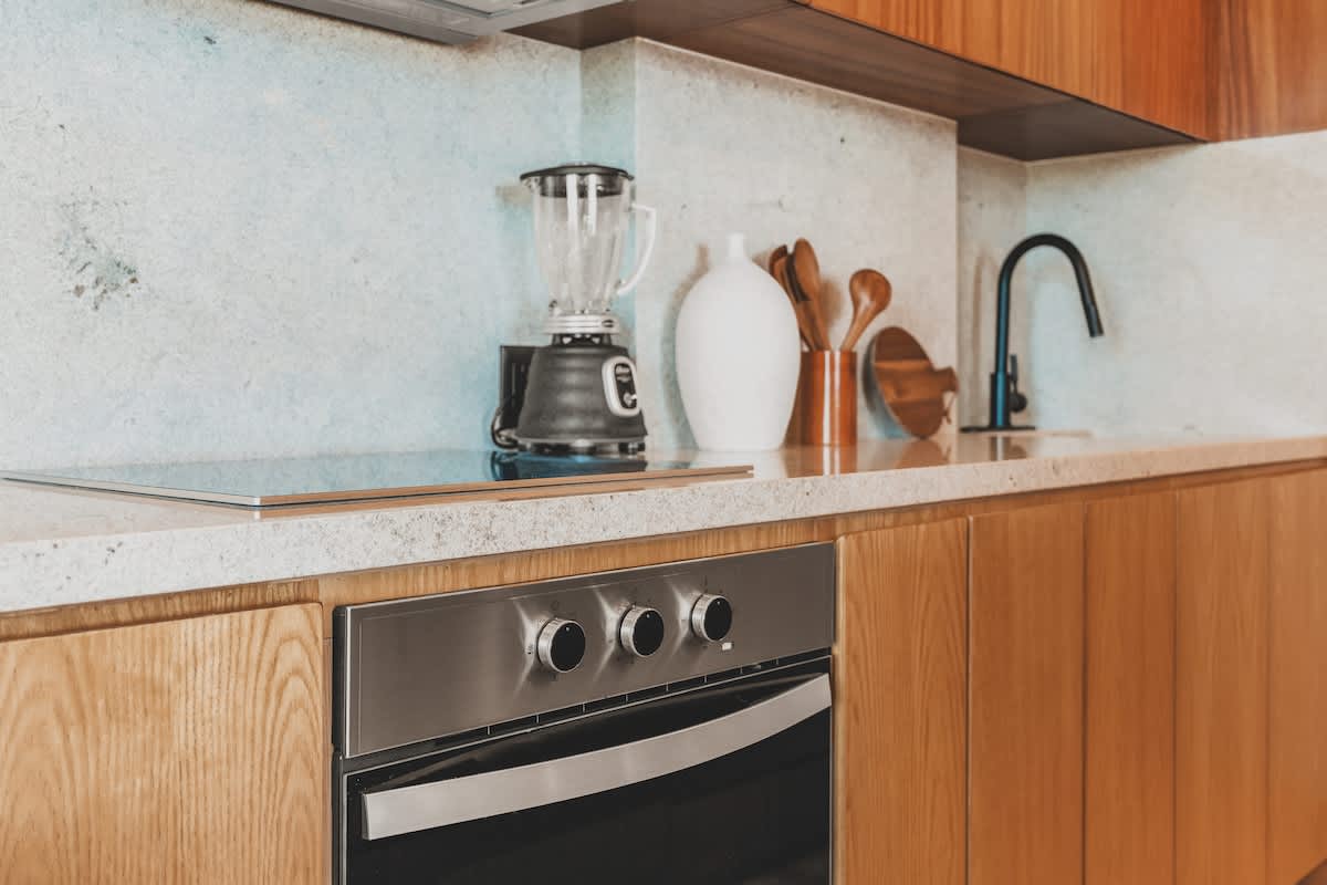 Modern kitchen counter with light-colored stone backsplash, featuring a blender, decorative vases with wooden utensils, and a built-in stainless steel oven below.