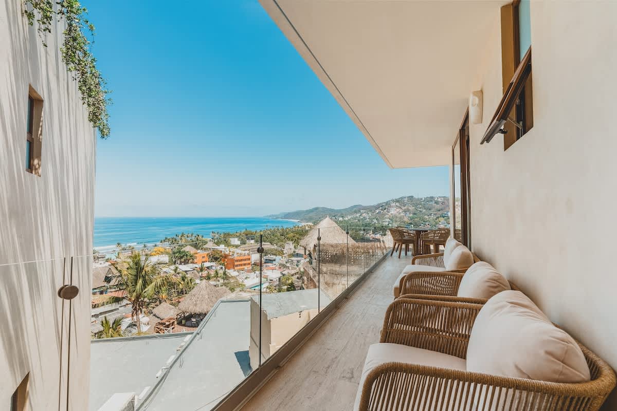 A spacious outdoor balcony offering panoramic ocean and town views, furnished with several woven lounge chairs with white cushions. A glass railing ensures an unobstructed vista under a clear blue sky.